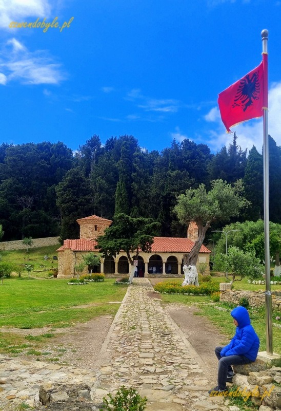 Albania, Zvernec. Brukowana kamieniami droga prowadzi do cerkwi. Po prawej maszt z powiewającą flagą Albanii. przy maszcie siedzi turysta w kurtce z kapturem. 20240504_125414-ink