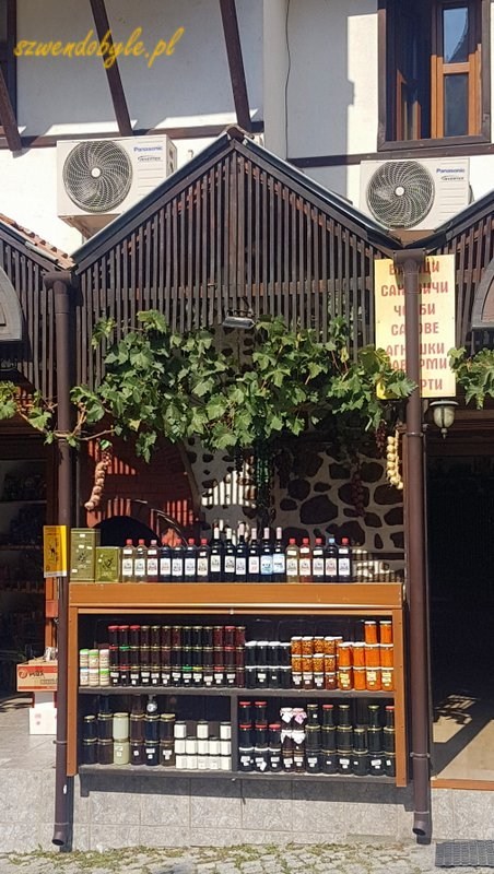 Bułgaria, Melnik (Мелник). Oferta sklepu z winem, regał przed sklepem. Butelki z winem i słoiki z przetworami stoją na półkach w pełnym słońcu. 20231003_140922-ink
