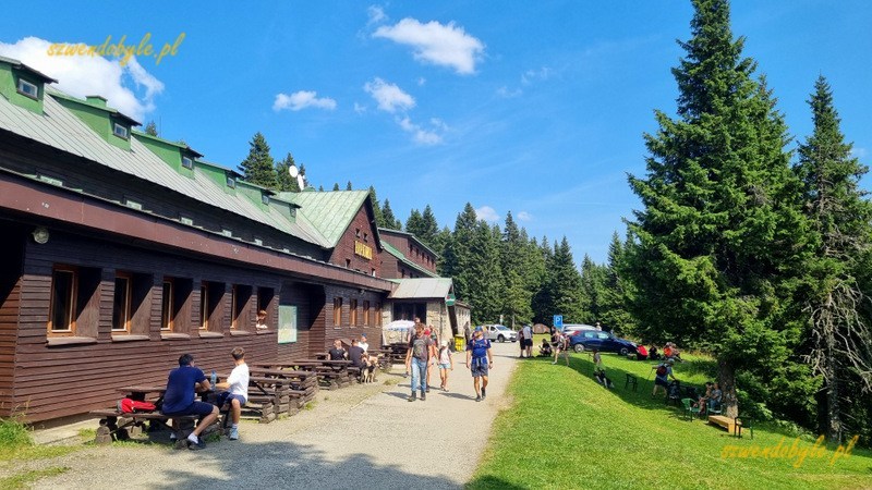 Czechy, widok na schronisko Barborka poniżej szczytu g&oacute;ry Pradziad. Przed drewnianym budynkiem stoją stoły i ławki,kilka os&oacute;b siedzi, inni spacerują. 20250815_142629-ink
