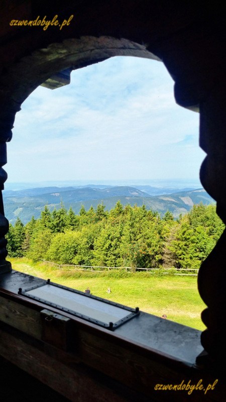 Czechy, Pustevny. Widok z okna dzwonnicy - punktu widokowego w kaplicy Cyryla i Metodego na g&oacute;rze Radho&scaron;ť (pol. Radhoszcz). Za oknem g&oacute;ry, po bokach obramowanie drewnianego otworu okiennego, rzeźbione belki z ciemnego drewna. Zdjęcie w pionie. 20250809_105304-ink