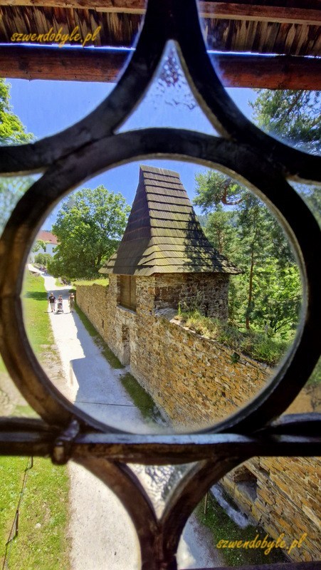Zamek Velhartice, Czechy. Widok na niewielką basztę przez kute kraty okna kawiarni nad bramą zamku. Basztę widać przez okrągły otwór krat. 20250620_130431-ink