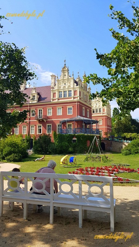 Bad Muskau, Neues Schloss / Mużaków. Nowy Zamek. Widok na boczne skrzydlo zamku,na pierwszym planie drewniana biała ławka, osoba dorosła i dziecko siedzą plecami do kamery. 20230614_162351-ink