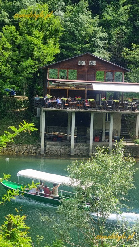 Czarnogóra, Rijeka Crnojevića, Widok z drugiego brzegu na restaurację z tarasem. Na wodzie płynie łódź z turystami. 20250504_141420-ink