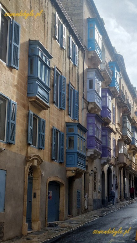 Maltańskie balkony, Valletta. Nagromadzenie kolorowych balkonów (niebieskie i fioletowe) na ścianach kamienic. Zdjęcie po deszczu, perspektywa ulicy.