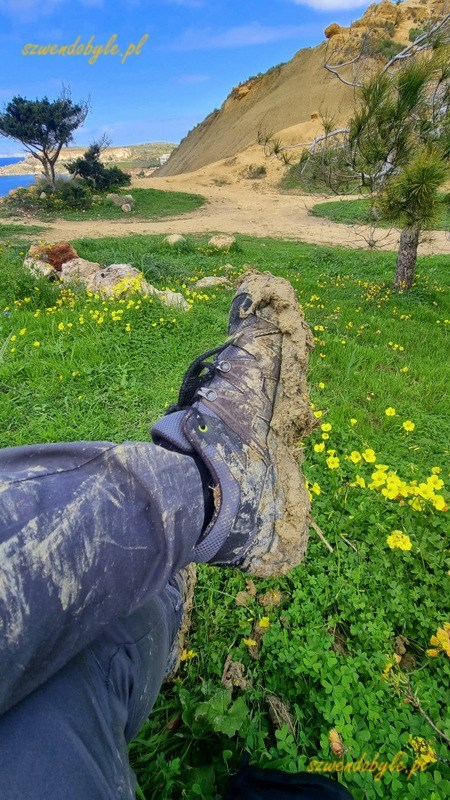Malta, pokryte błotem buty na tle zielonej trawy z żółtymi kwiatami polnymi. W oddali pagórek widokowy. 20250308_123408-ink