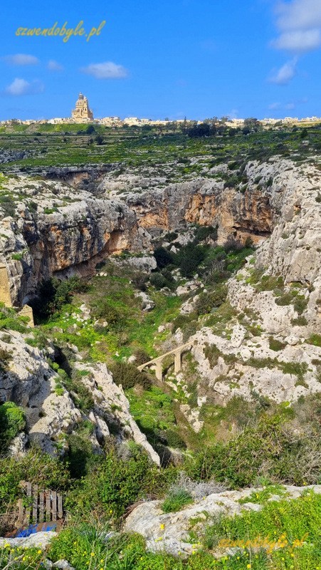 Malta, wyspa Gozo. Widok z drogi w kierunku miasteczka Ix-Xewkija. Nad miastem góruje kopuła kościelna. U dołu wąwóz pomiędzy skałami. 20250303_123321-ink