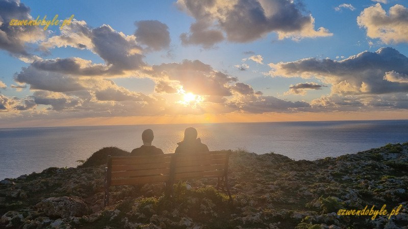 Malta, na pierwszym planie ławeczka i cień dwóch osób oglądających zachód słońca z klifów Dingli. 