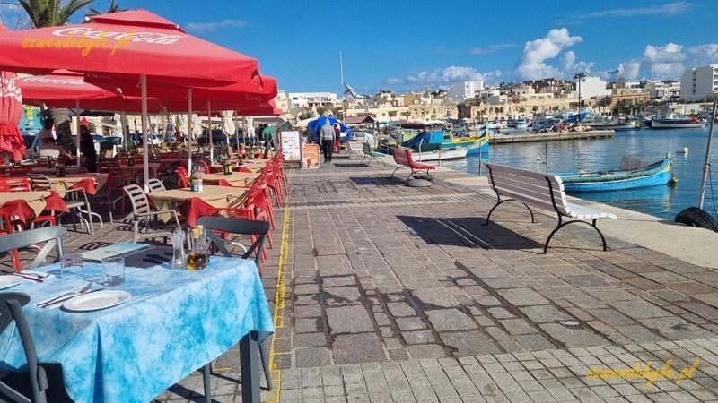 Malta, Marsaxlokk. Widok wzdłuż nabrzeża w porcie - po lewej stoliki i czerwone parasole restauracji, po prawej brzeg morza.