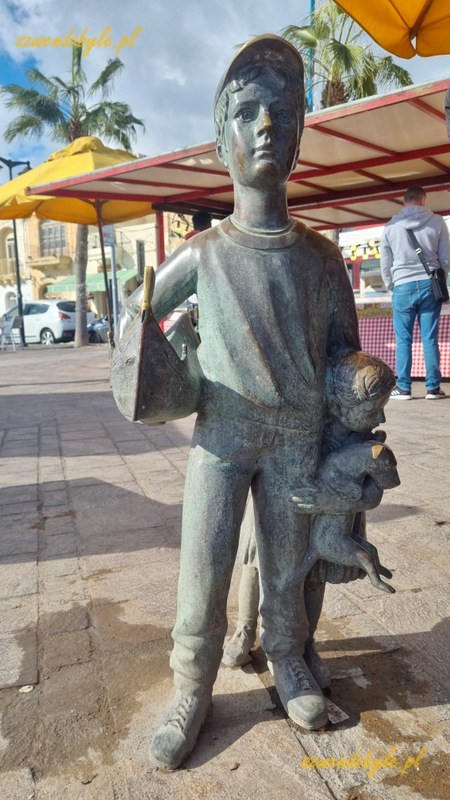 Malta, Marsaxlokk. Pomnik w porcie - figura chłopca, syna rybaka witającego ojca. Pod pachą trzyma model łodzi. Zza chłopca wychyla się jego siostra.