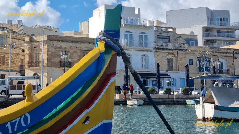 Malta, Marsaxlokk. Widok z łodzi na miasteczko. Na pierwszym planie fragment kolorowej łodzi.