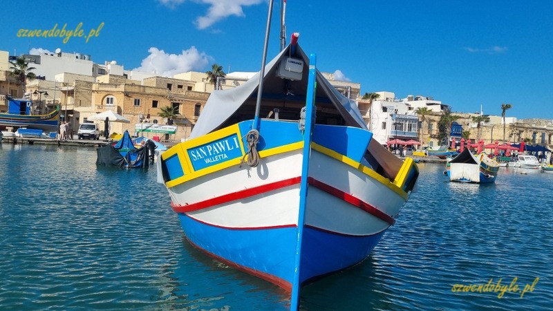 Malta, Marsaxlokk. Kolorowa łódź zacumowana w porcie. Zdjęcie wykonane z wody, widać budynki miasteczka.