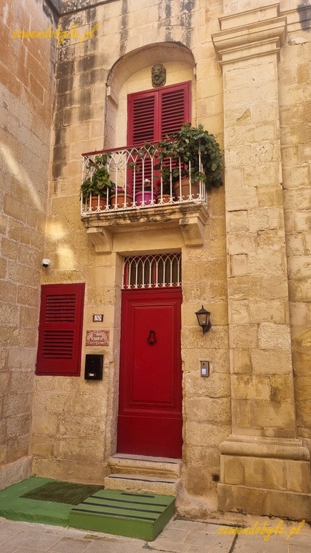 Mdina, niewielki kamienny zaułek. W rogu czerwone drzwi, ponad nimi niewielki balkon.