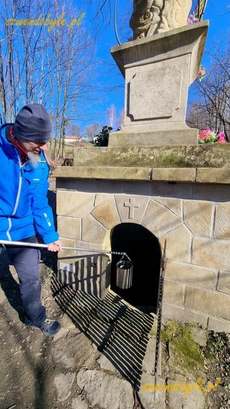 Małopolska, Bocheniec. Kamienna rzeźba św. Anny Samotrzeć. Powyżej rzeźby niewielki daszek. W postumencie są kratowane drzi do źr&oacute;dełka, wodę można nabrać (jak ze studni) niewielkim metalowym wiadrem zawieszonym na metalowym kiju. 20260307_091506-ink