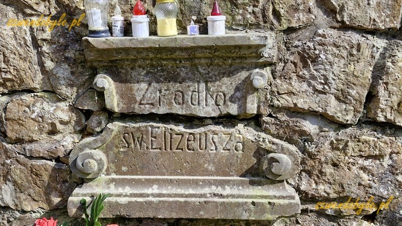 Małopolska, kamienna tablica z napisem "Źr&oacute;dło św. Elizeusza". Widoczne poziome pęknięcie tablicy. Powyżej tablicy stoją znicze. 20260315_120338-ink