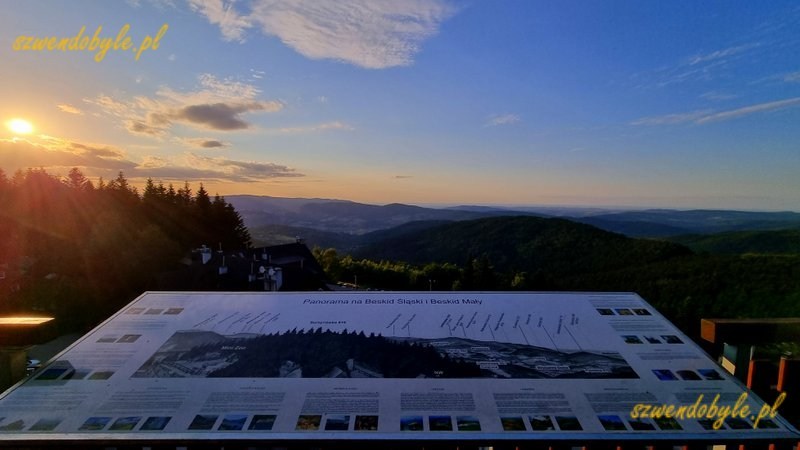 Małopolska,Stryszawa - widoki z wieży widokowej Beskidzki Raj. Na pierwszym planie tablica z opisem "Panorama na Beskid Śląski i Beskid Mały". Po lewej zachodzące słońce.20260704_195245-ink