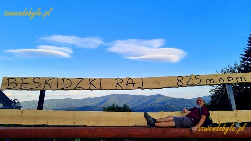 Małopolska,Stryszawa - pod wieżą widokową Beskidzki Raj, na poziomie parkingu, znajduje się gigantyczna ławka - pomiędzy deskami oparcia widać panoramę na Gorce. Na górnej desce oparcia napis "Beskidzki Raj 825 m n.m.p.". 20260704_195949-ink