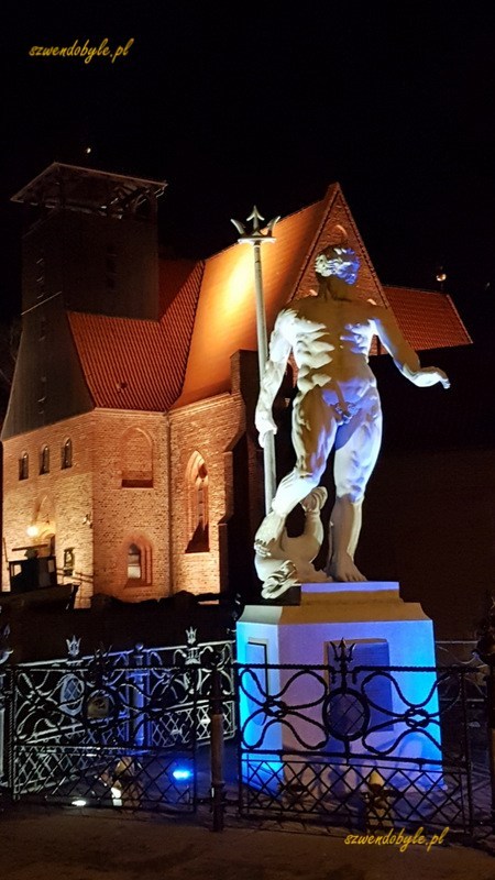 Hel, zdjęcie nocne. Oświetlkony pomnik Neptuna na tle starego kościoła ewangelickiego (obecnie muzeum rybołówstwa). Budynek kościoła oświetlony. Widać niewysokie ogrodzenie pomnika.