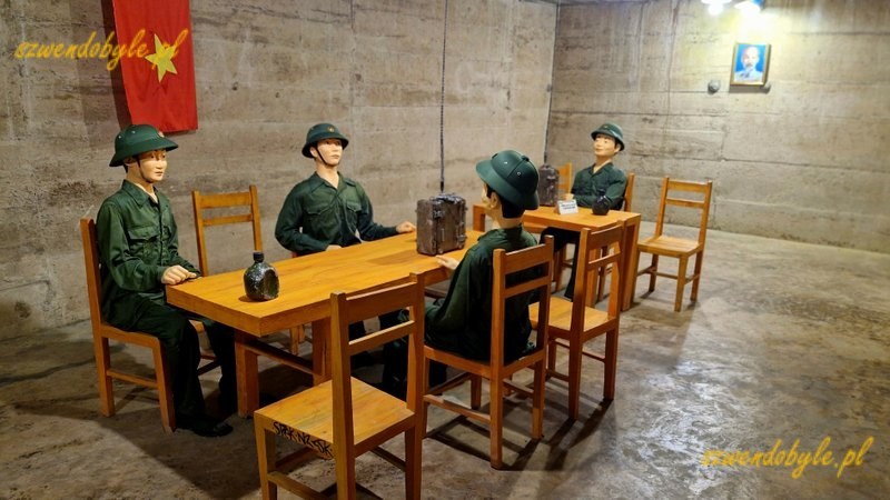 Wietnam, wyspa Cát Bà, jaskinia, w której mieścił się szpital wojskowy, czyli Military Hospital Cave. Sala konferencyjna, stół i krzesła, 4 manekiny w mundurach siedzą na krzesłach wokół stołu. Na jednej ścianie wisi flaga Wietnamu, na drugiej ścianie niewielki portret Ho Chi Minha. 20251124_114426-ink