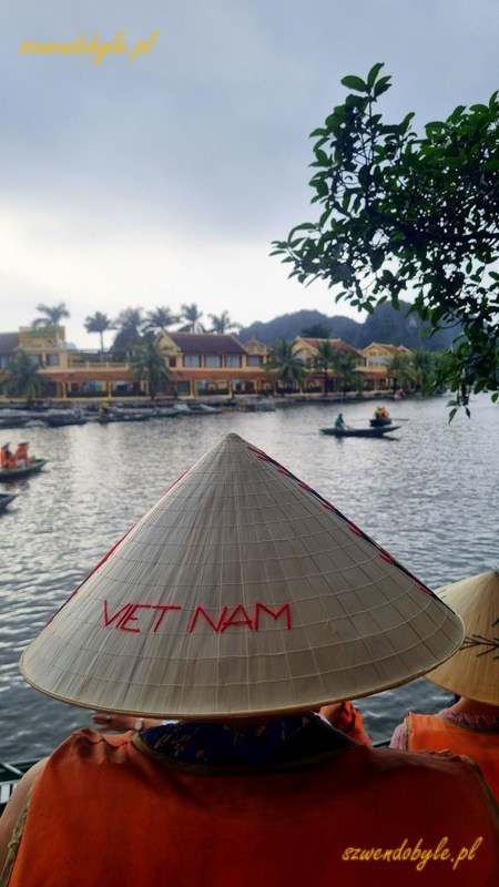 Tam Coc, Vietnam. Na pierwszym planie postać w tradycyjnym stożkowym kapeluszu z czerwonym napisem VIET NAM. 20251110_150349-ink