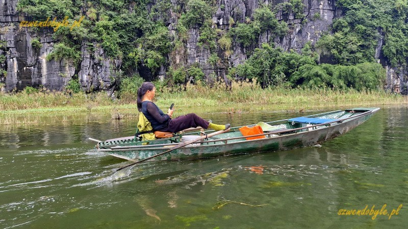 Tam Coc, Vietnam. Ł&oacute;dź bez pasażer&oacute;w na rzece Ngo Dong,wioślarka macha wiosłami używając st&oacute;p i jednocześnie korzysta ze smartfona. 20251110_162756-ink