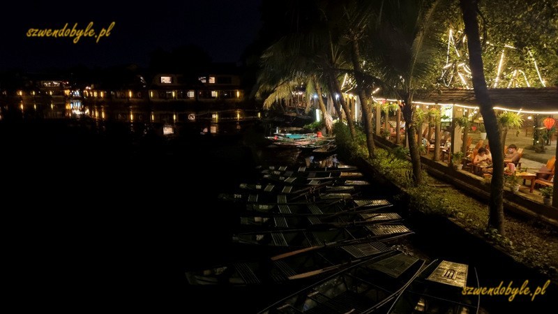 Tam Coc, Vietnam. Widok po zmroku z mostku na terenie miasteczka - na pierwszym planie ł&oacute;dki przycumowane przy brzegu, na brzegu oświetlony bar. 20251110_182505js-ink