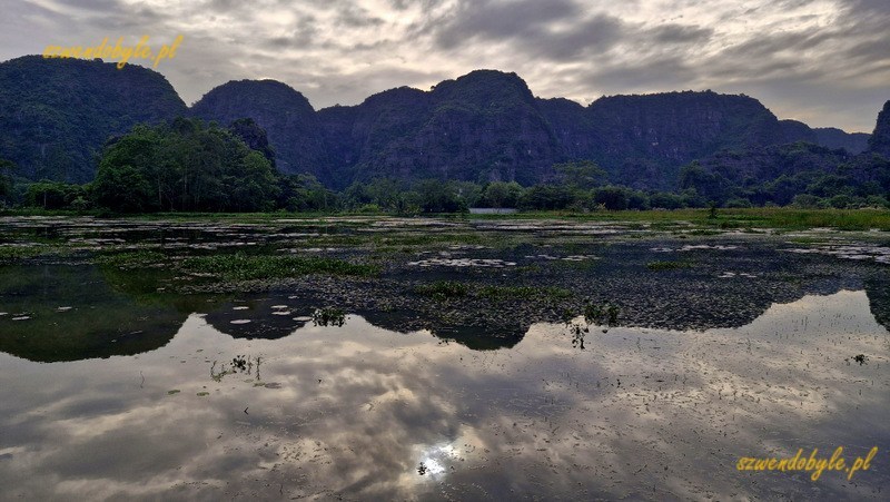 Tam Coc, Vietnam. Spokojna rzeka Ngo Dong bez łodzi. Pochmurne niebo i g&oacute;ry odbijają się w wodzie. 20251111_152930-ink