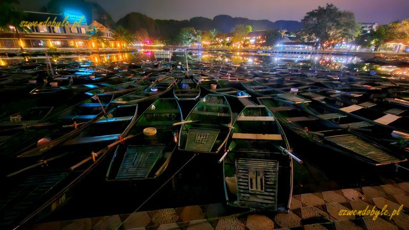 Tam Coc, Vietnam. Wiele łodzi przycumowano nieopodal przystani w miasteczku. Zdjęcie wykonane po zmroku,na drugim brzegu widać wiele kolorowych światełek z bar&oacute;w. 20251111_201604-ink