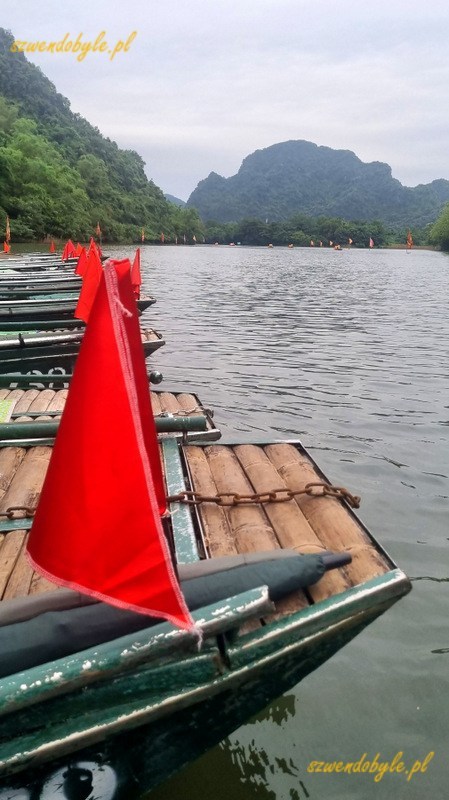 Tr&agrave;ng An, dzioby łodzi sampan z czerwonymi chorągiewkami. Łodzie czekają na turyst&oacute;w. 20251112_084404-ink